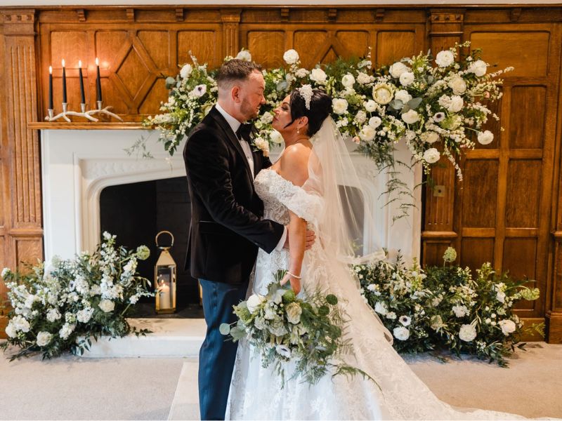 Micro Weddings For Meaningful Moments - Bride and groom looking at each standing in front of white fireplace surrounded by white and green flowers